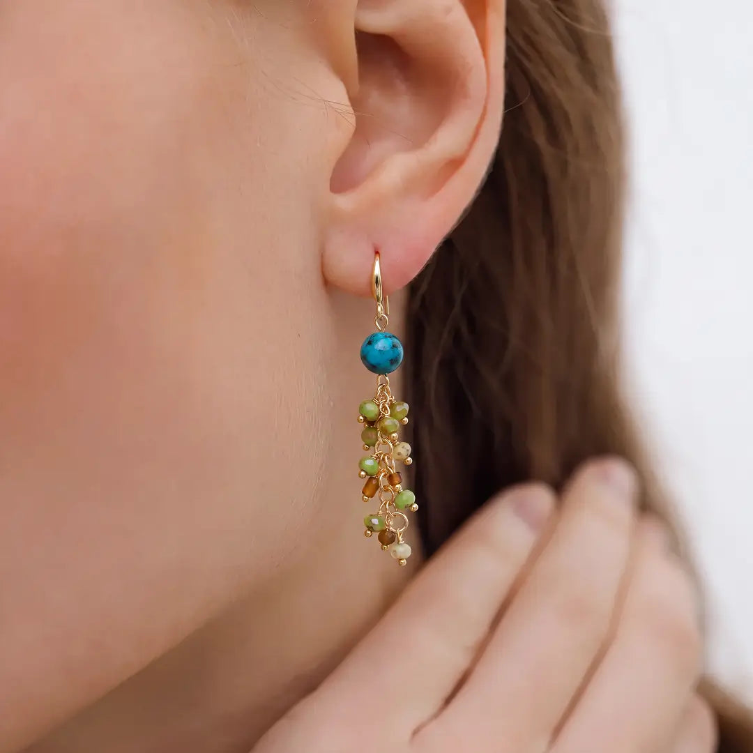 A close-up of a model's ear wearing a delicate gold dangle earring with a turquoise blue bead and green and gold bead accents.