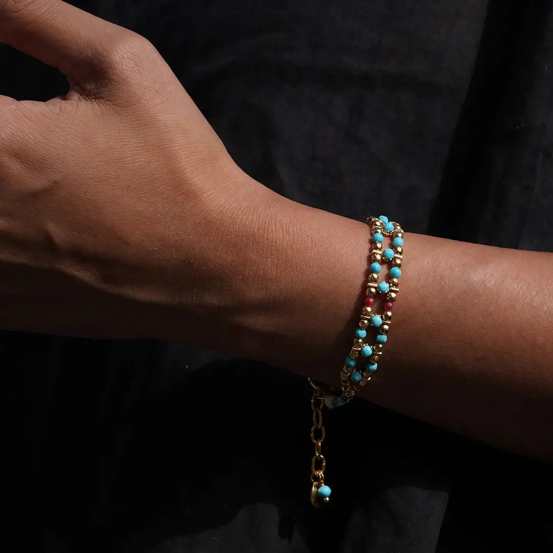 A close-up of a person's wrist wearing a delicate beaded bracelet with turquoise blue beads, gold-toned metal links, and small red beads, set against a dark background.