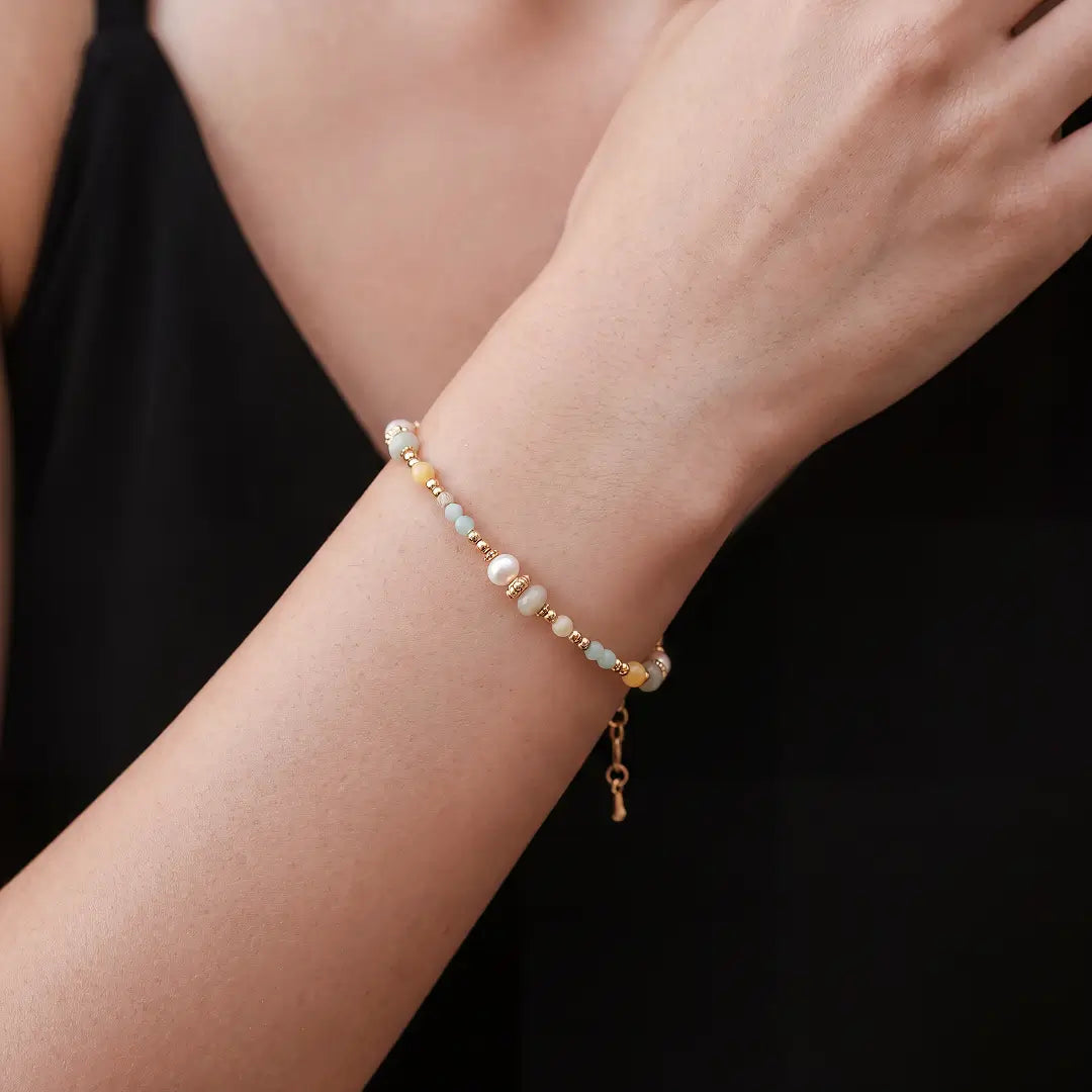A delicate beaded bracelet with white pearls, light blue beads, and gold-toned spacer beads, shown worn on a person's wrist against a black top.