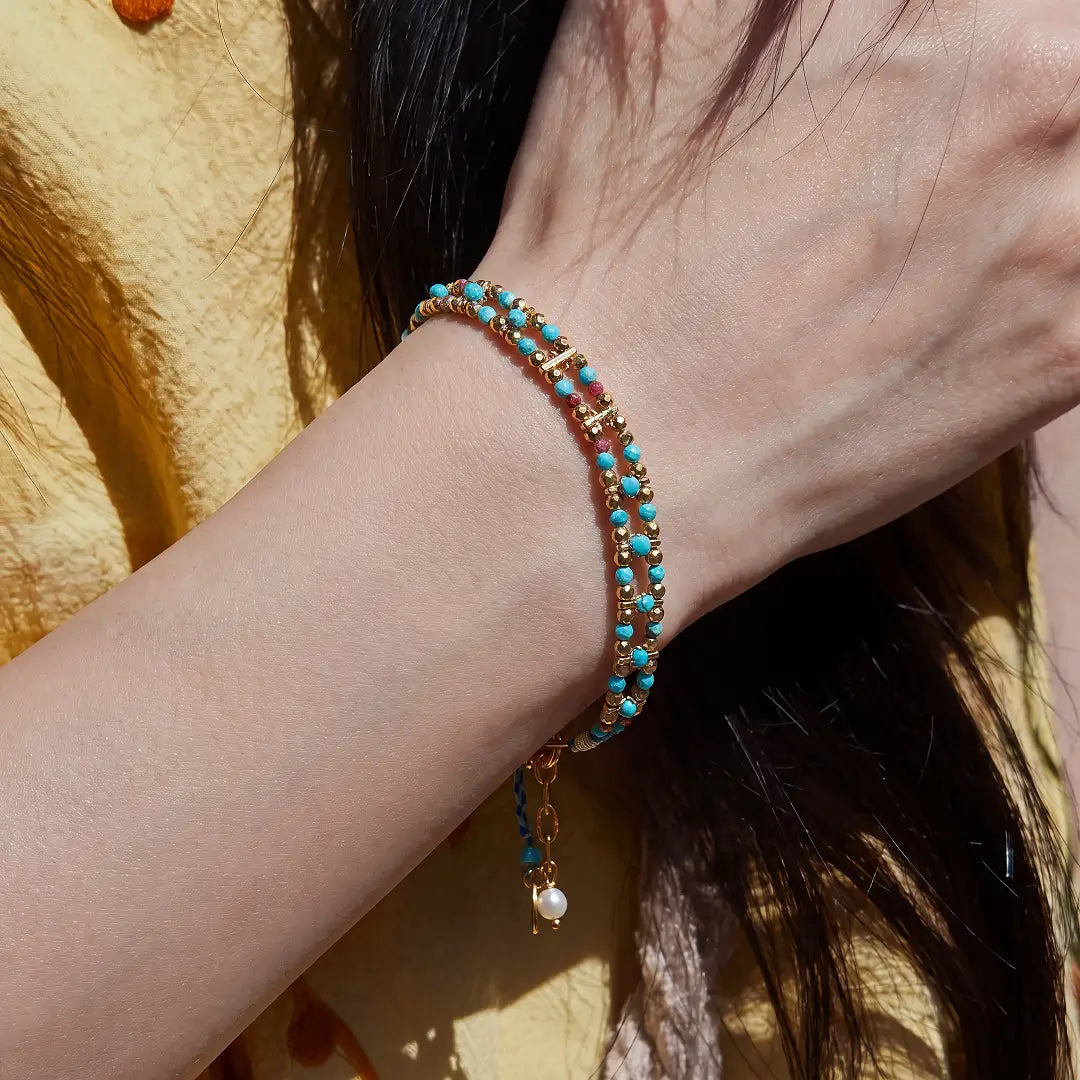 A close-up of a person's wrist wearing a double-strand beaded bracelet with turquoise blue beads, gold-toned spacer details, and a small dangling pearl charm.