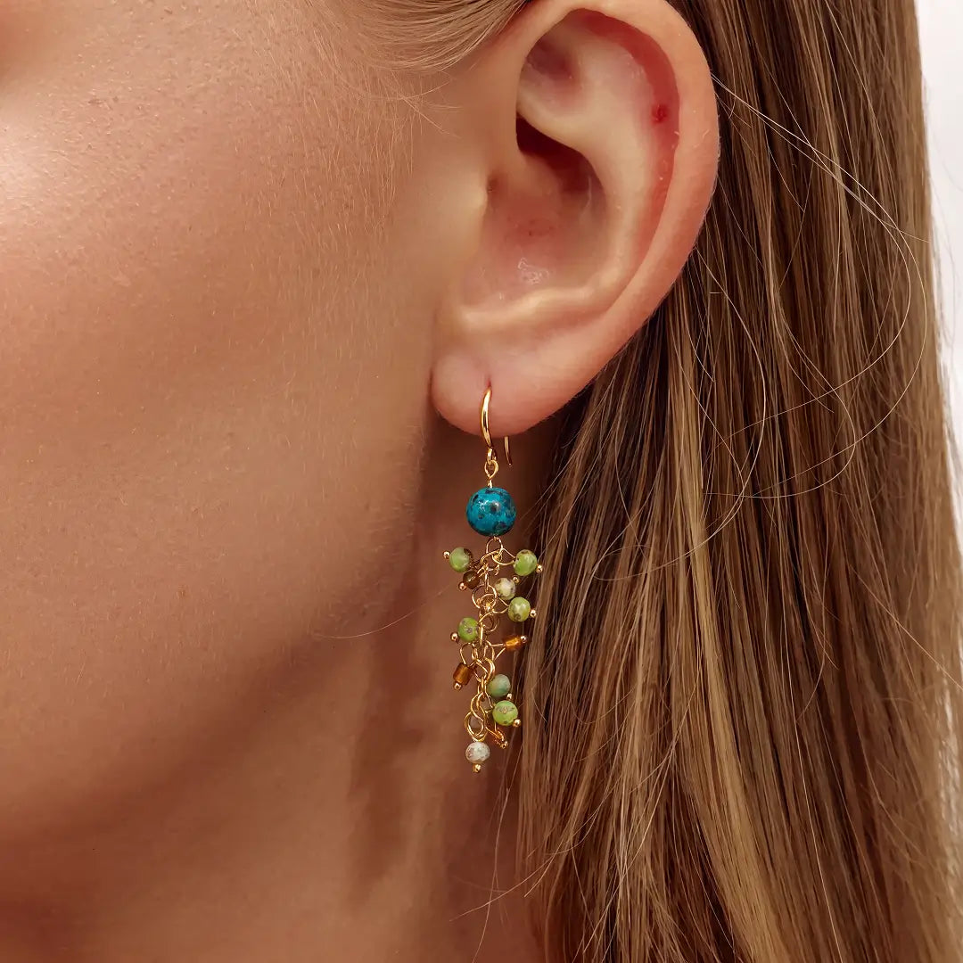 A close-up of a woman's ear wearing a dangling gold earring with a turquoise blue bead and green bead accents.